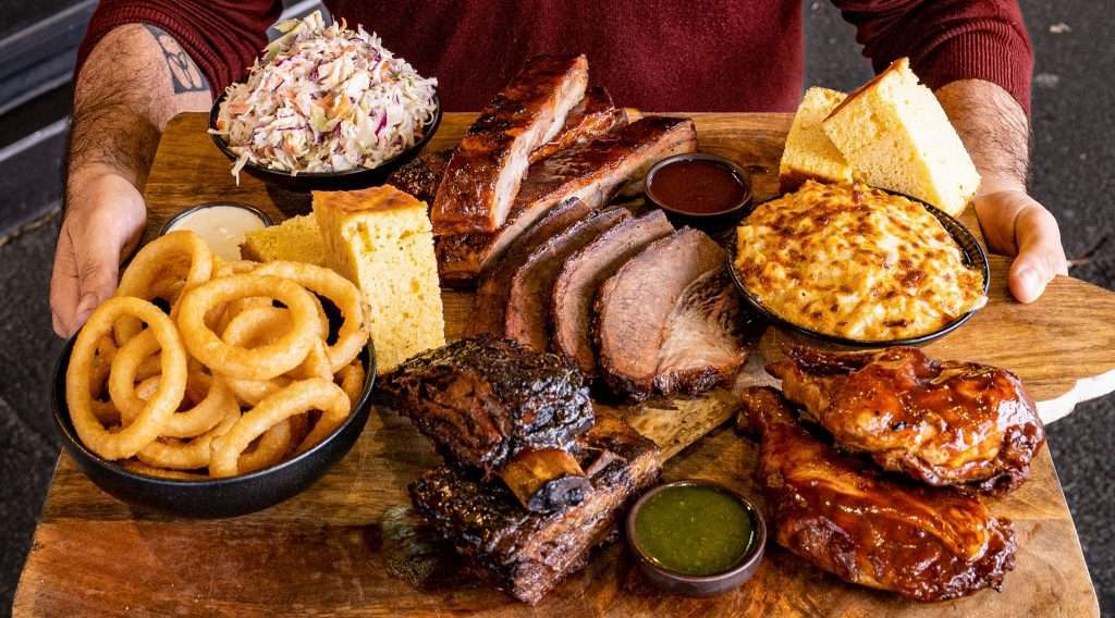 Person holding a large board of assorted smoked meats and sides