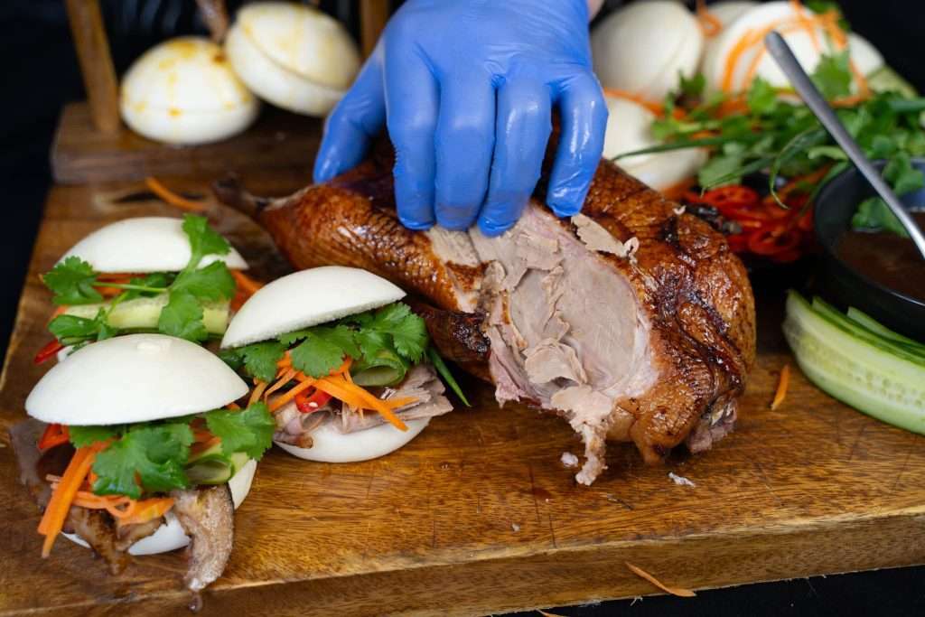 Gloved hand carving smoked duck with tongs beside bao buns