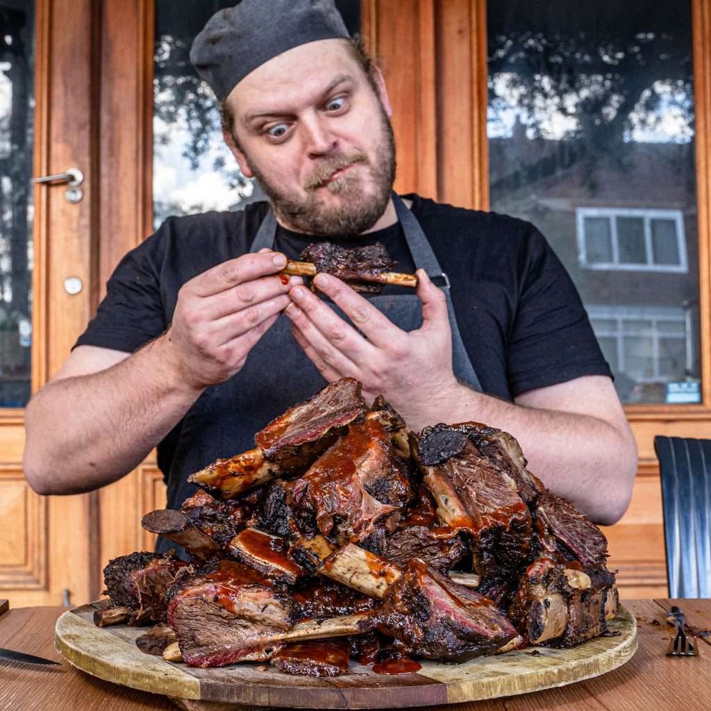Chef eating a rib beside a large pile of smoked beef ribs